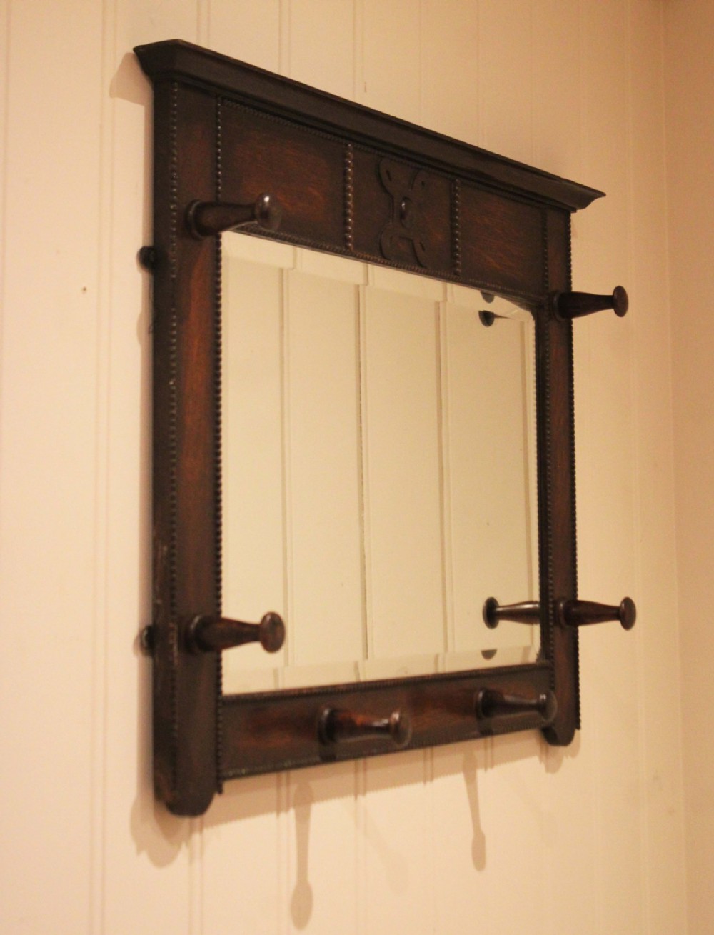 A dark oak rectangular wall mirror featuring six turned wooden coat pegs framing the glass plate, decorated with beaded trim and a carved central boss on the top rail. View VI