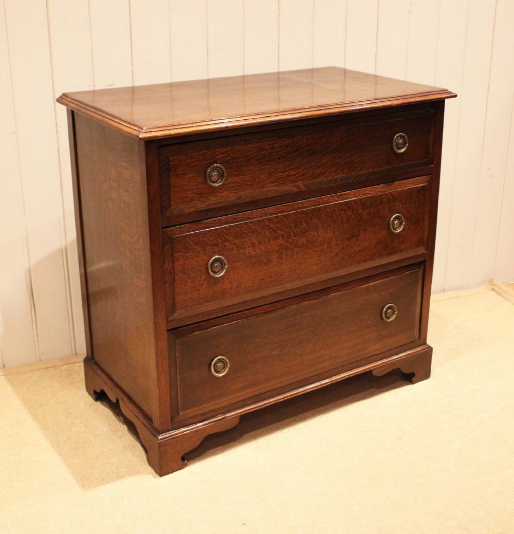 A solid oak chest of drawers featuring three full-width drawers with circular brass drop ring handles and bracket feet. View V