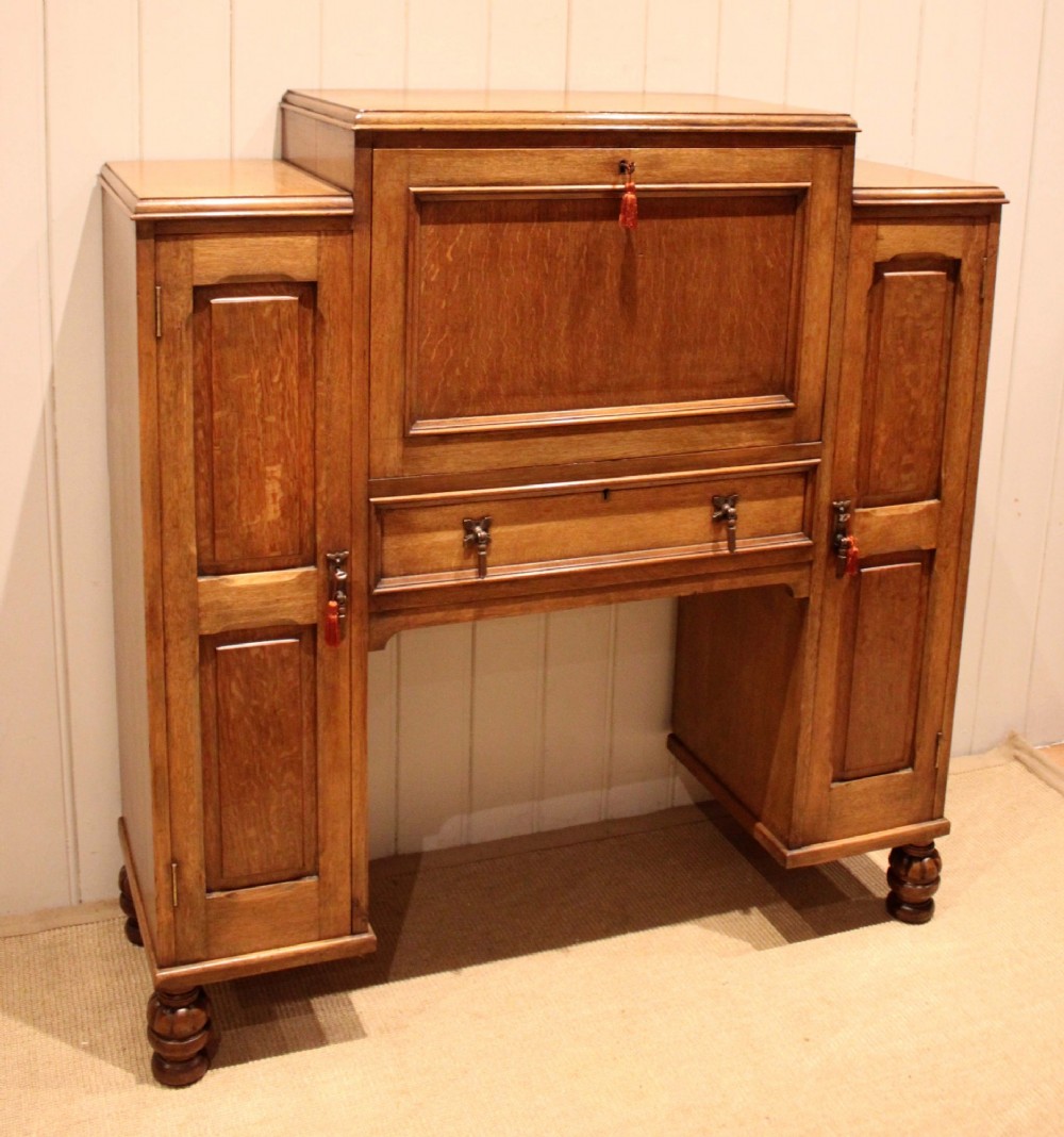 An antique oak bureau cabinet featuring a central fall-front writing compartment above a single drawer, flanked by two side cupboards with panelled doors and raised on turned feet. View II