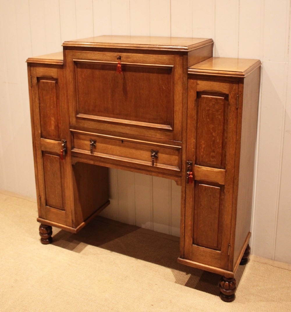 An antique oak bureau cabinet featuring a central fall-front writing compartment above a single drawer, flanked by two side cupboards with panelled doors and raised on turned feet. View III