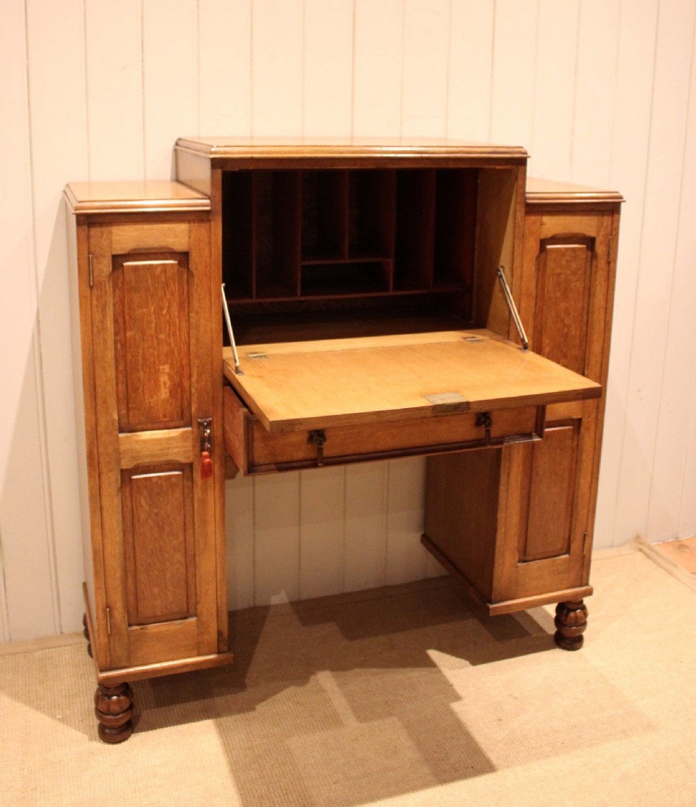 An antique oak bureau cabinet featuring a central fall-front writing compartment above a single drawer, flanked by two side cupboards with panelled doors and raised on turned feet. View V