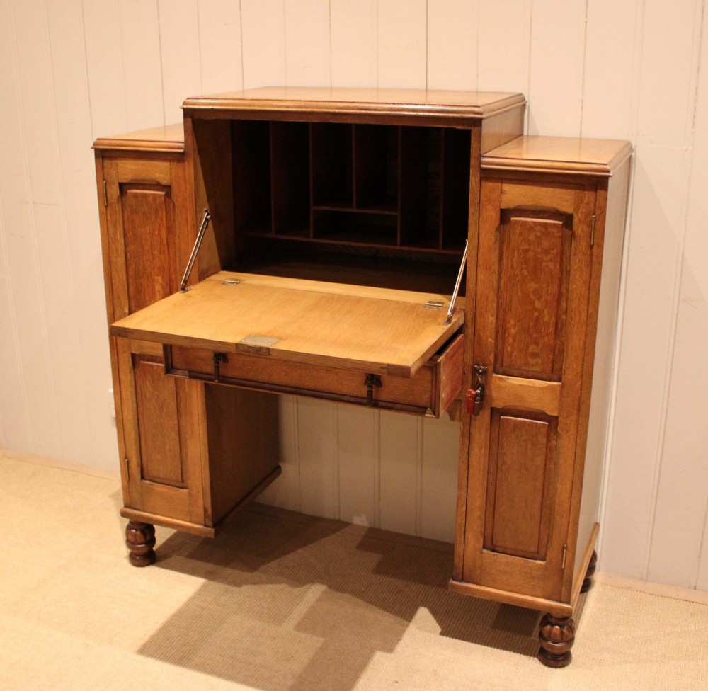 An antique oak bureau cabinet featuring a central fall-front writing compartment above a single drawer, flanked by two side cupboards with panelled doors and raised on turned feet. View VI
