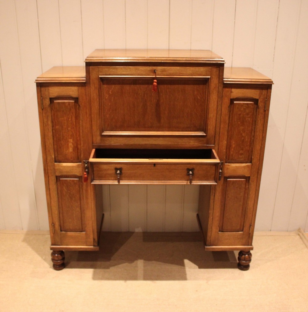 An antique oak bureau cabinet featuring a central fall-front writing compartment above a single drawer, flanked by two side cupboards with panelled doors and raised on turned feet. View VII