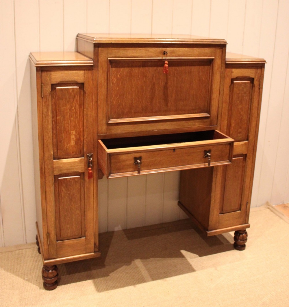 An antique oak bureau cabinet featuring a central fall-front writing compartment above a single drawer, flanked by two side cupboards with panelled doors and raised on turned feet. View VIII