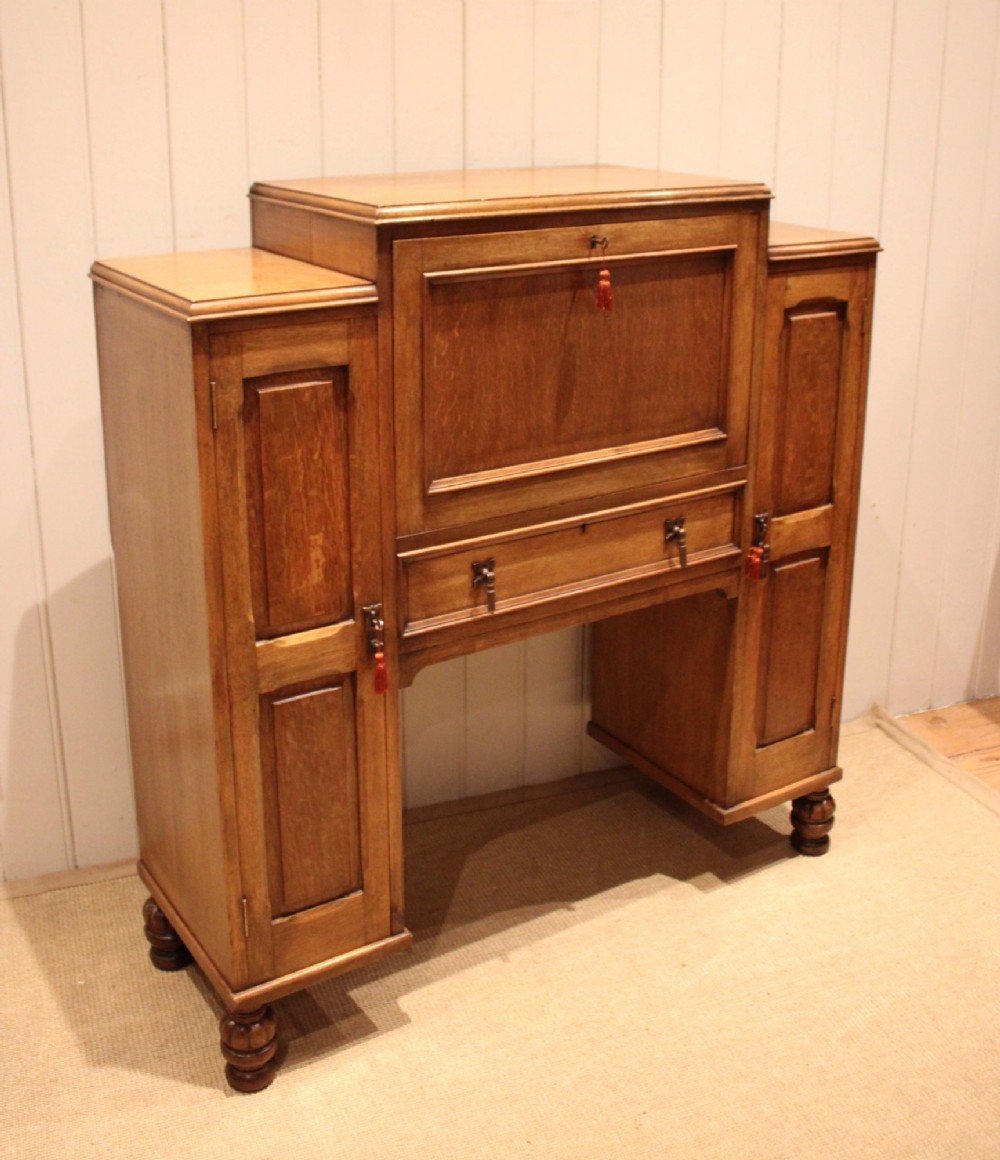 An antique oak bureau cabinet featuring a central fall-front writing compartment above a single drawer, flanked by two side cupboards with panelled doors and raised on turned feet. View X