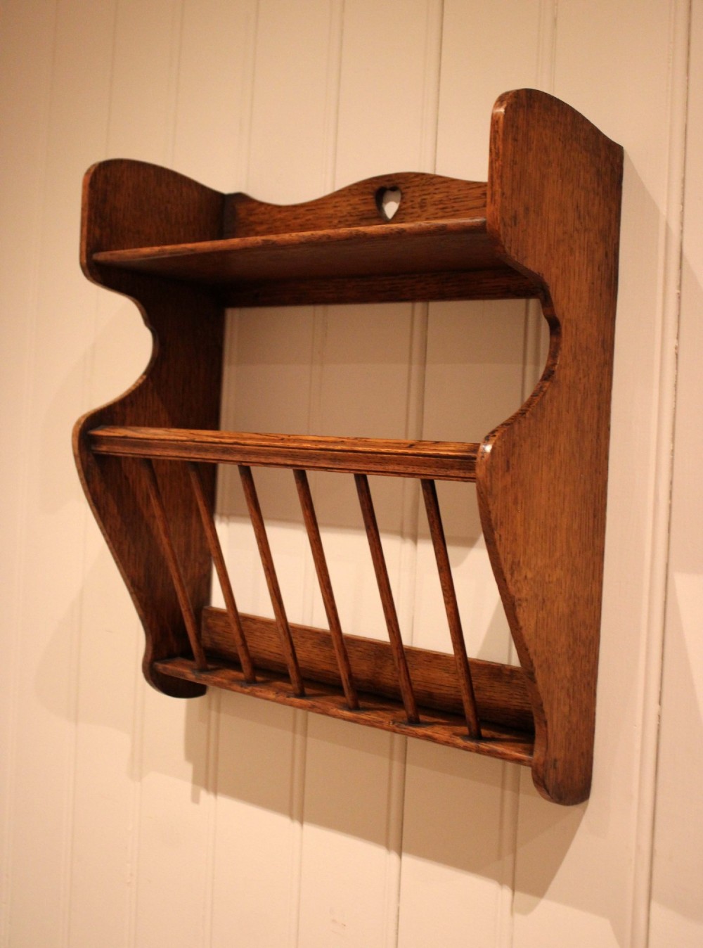An Arts and Crafts solid oak wall shelf featuring a top shelf ledge with a pierced heart cutout and a lower magazine rack with vertical wooden spindles.