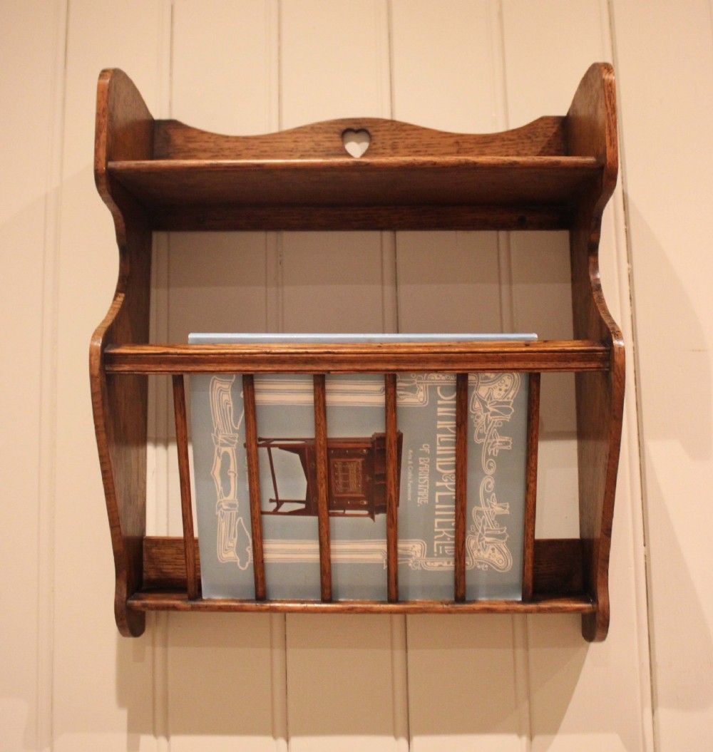 An Arts and Crafts solid oak wall shelf featuring a top shelf ledge with a pierced heart cutout and a lower magazine rack with vertical wooden spindles. View VI