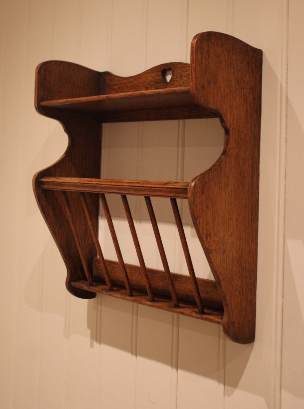 An Arts and Crafts solid oak wall shelf featuring a top shelf ledge with a pierced heart cutout and a lower magazine rack with vertical wooden spindles. View VII