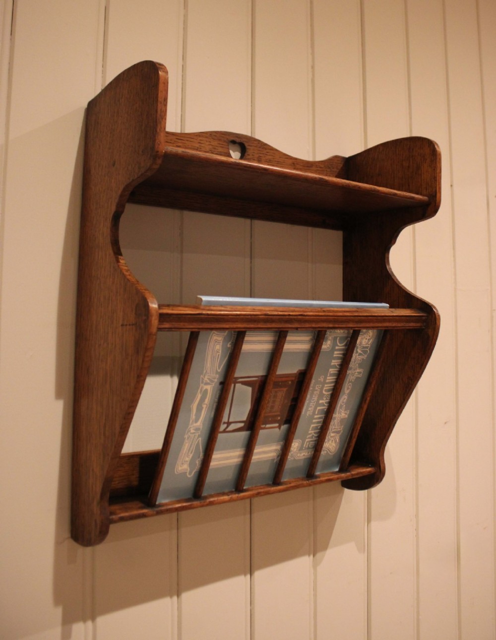 An Arts and Crafts solid oak wall shelf featuring a top shelf ledge with a pierced heart cutout and a lower magazine rack with vertical wooden spindles. View IX