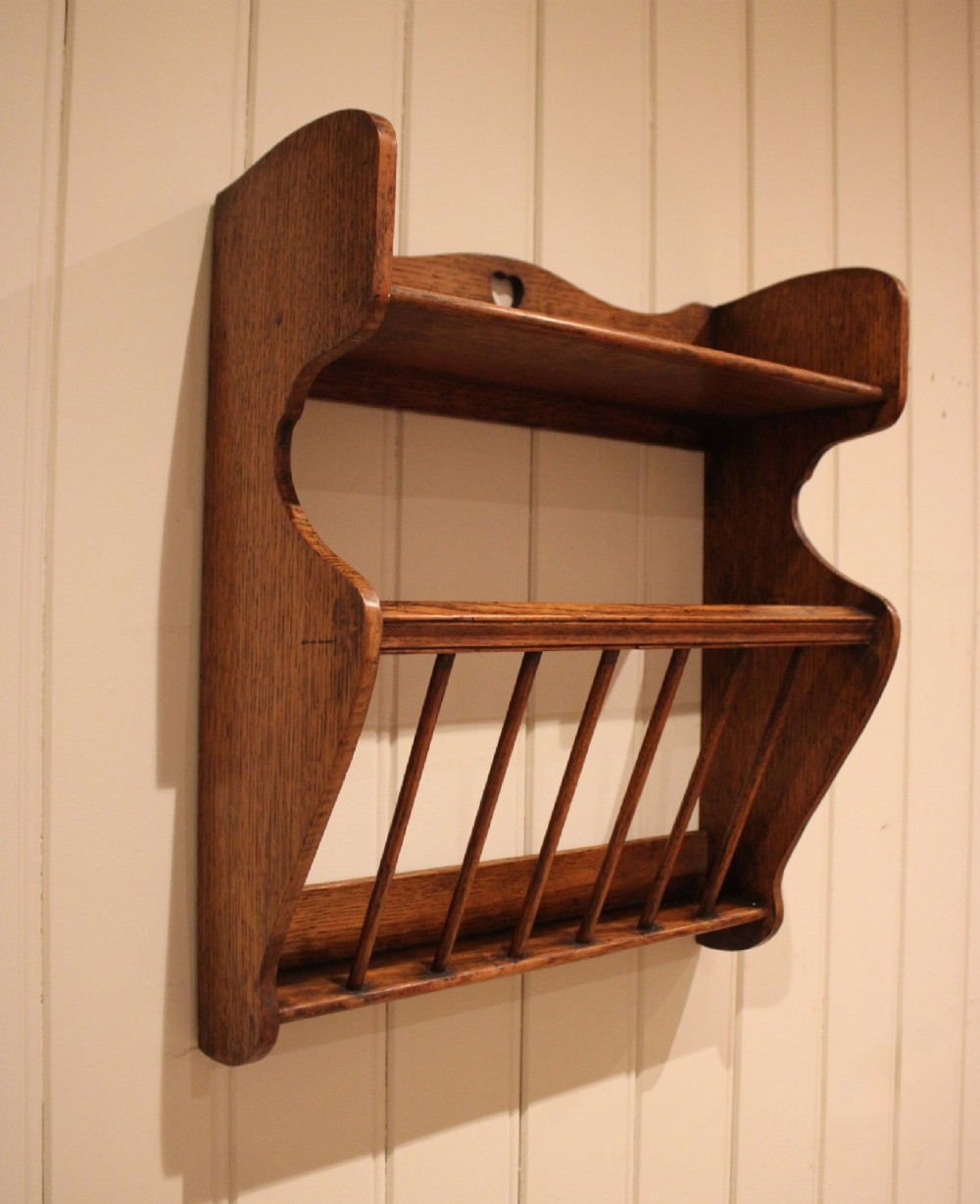 An Arts and Crafts solid oak wall shelf featuring a top shelf ledge with a pierced heart cutout and a lower magazine rack with vertical wooden spindles. View X