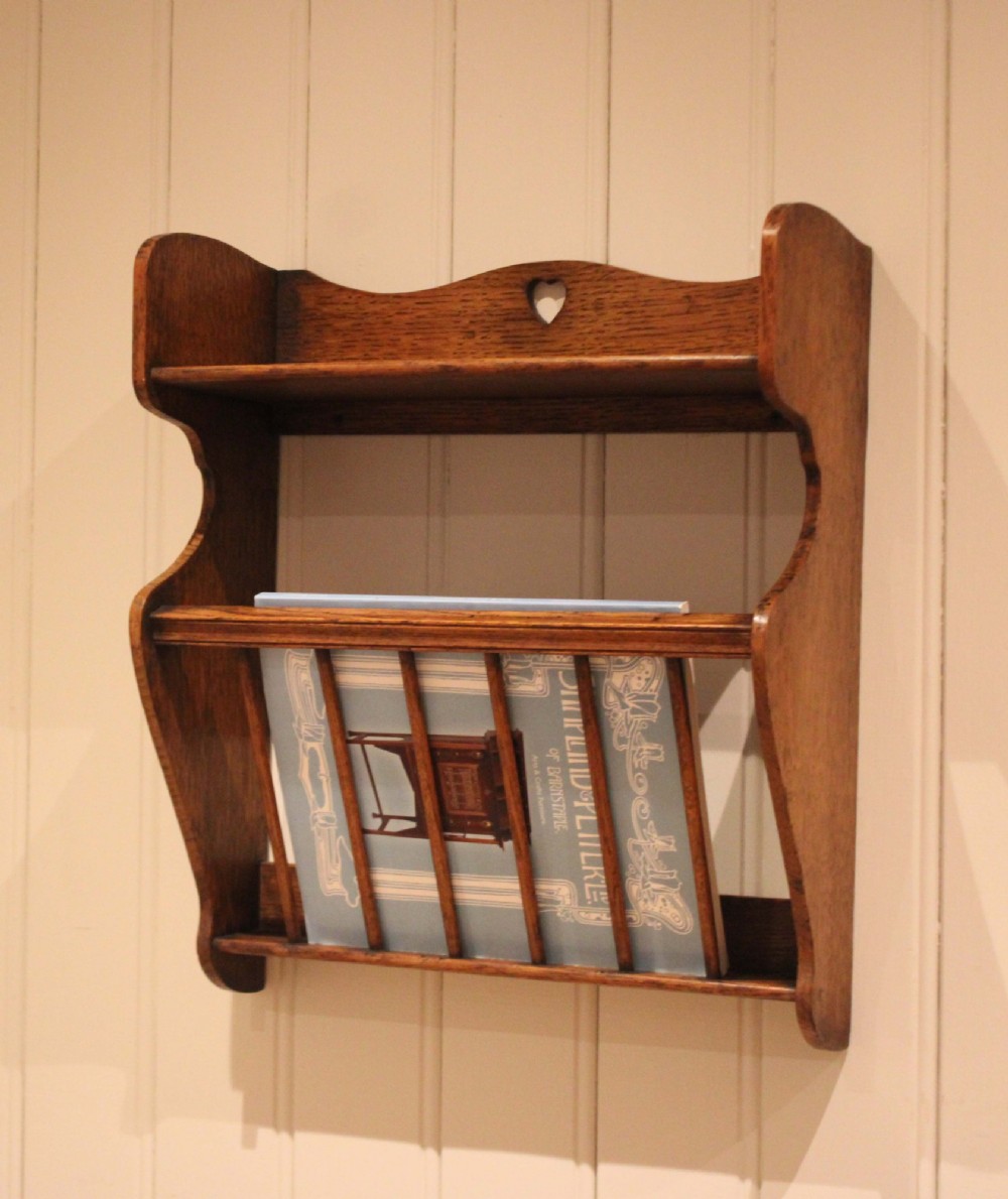 An Arts and Crafts solid oak wall shelf featuring a top shelf ledge with a pierced heart cutout and a lower magazine rack with vertical wooden spindles. View XII