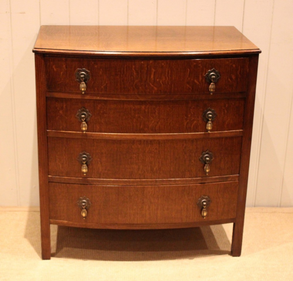 A dark oak chest of drawers featuring a curved bowfront facade and four graduated drawers with antique brass pendant handles.