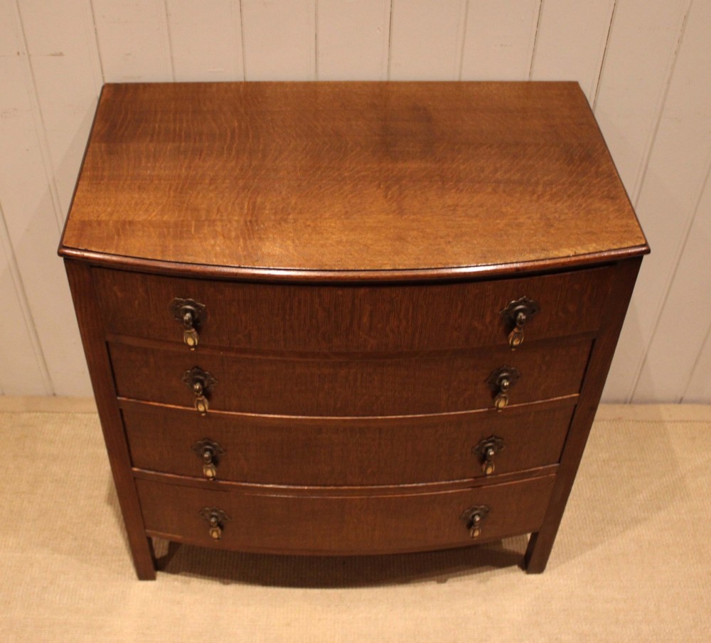 A dark oak chest of drawers featuring a curved bowfront facade and four graduated drawers with antique brass pendant handles. View IV