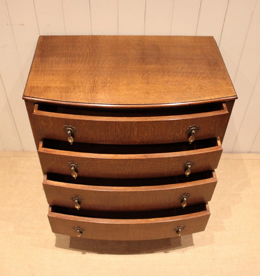 A dark oak chest of drawers featuring a curved bowfront facade and four graduated drawers with antique brass pendant handles. View V