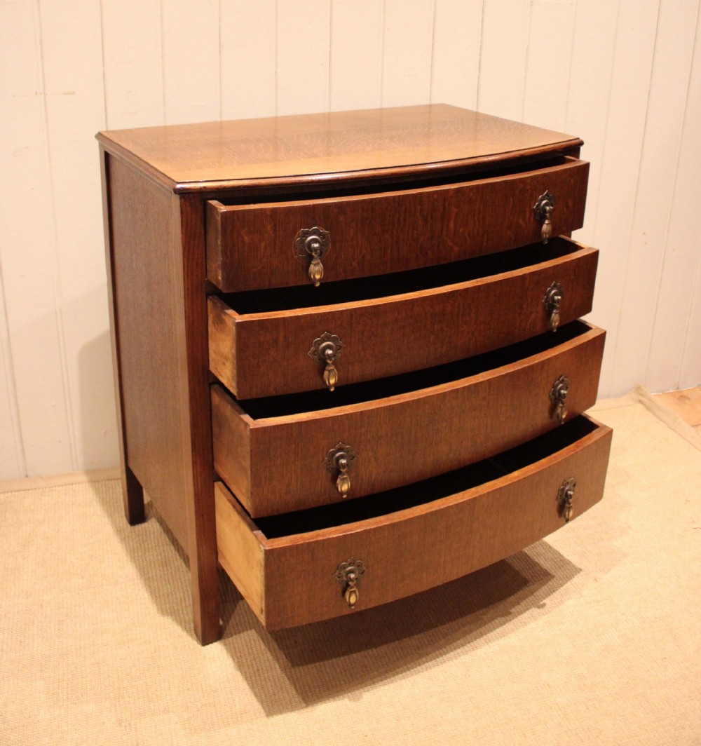 A dark oak chest of drawers featuring a curved bowfront facade and four graduated drawers with antique brass pendant handles. View VI