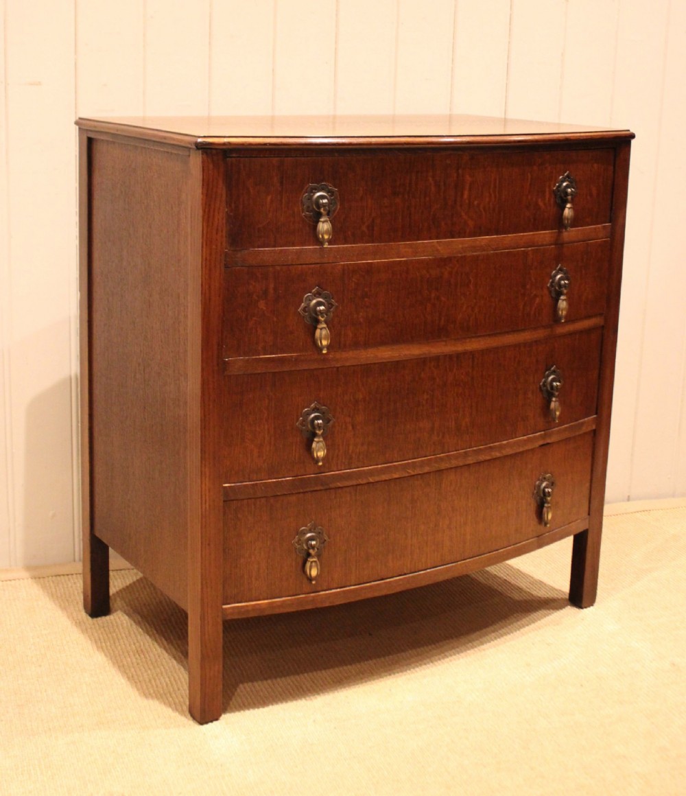 A dark oak chest of drawers featuring a curved bowfront facade and four graduated drawers with antique brass pendant handles. View VII