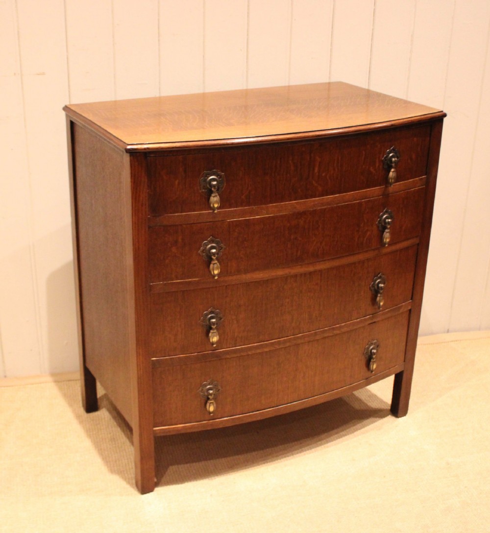 A dark oak chest of drawers featuring a curved bowfront facade and four graduated drawers with antique brass pendant handles. View IX