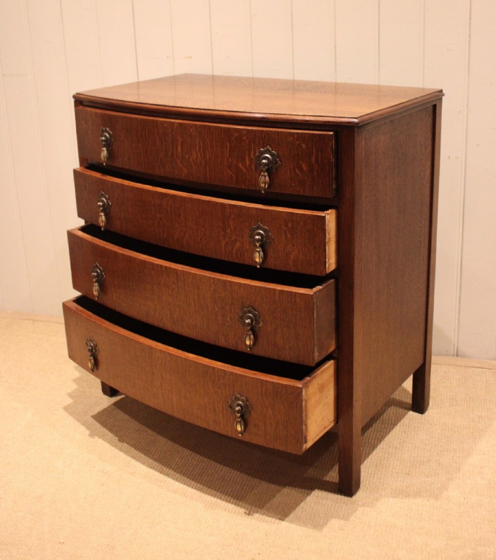 A dark oak chest of drawers featuring a curved bowfront facade and four graduated drawers with antique brass pendant handles. View X
