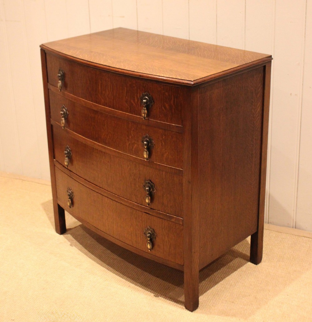 A dark oak chest of drawers featuring a curved bowfront facade and four graduated drawers with antique brass pendant handles. View XI