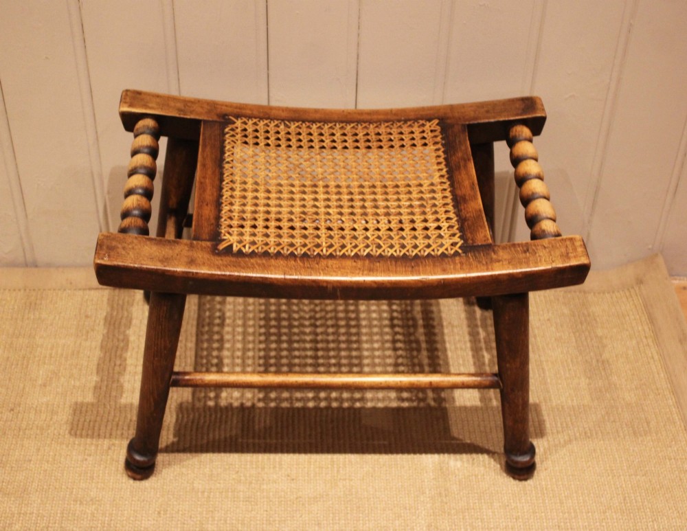 An antique warm brown beechwood stool featuring a woven cane seat, bobbin-turned side handles, and splayed turned legs.