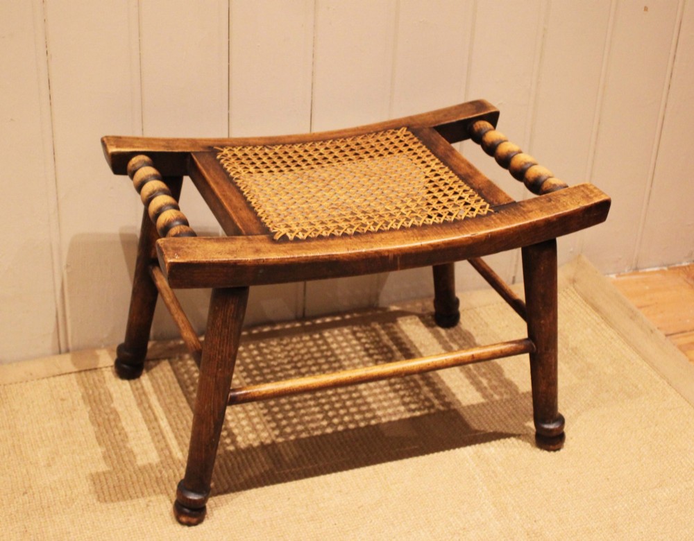 An antique warm brown beechwood stool featuring a woven cane seat, bobbin-turned side handles, and splayed turned legs. View II