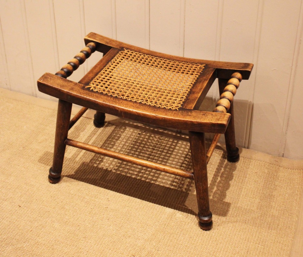 An antique warm brown beechwood stool featuring a woven cane seat, bobbin-turned side handles, and splayed turned legs. View III