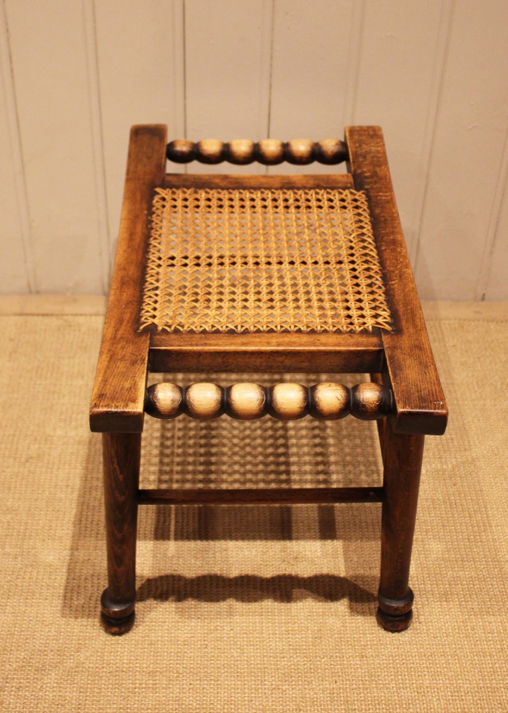 An antique warm brown beechwood stool featuring a woven cane seat, bobbin-turned side handles, and splayed turned legs. View V