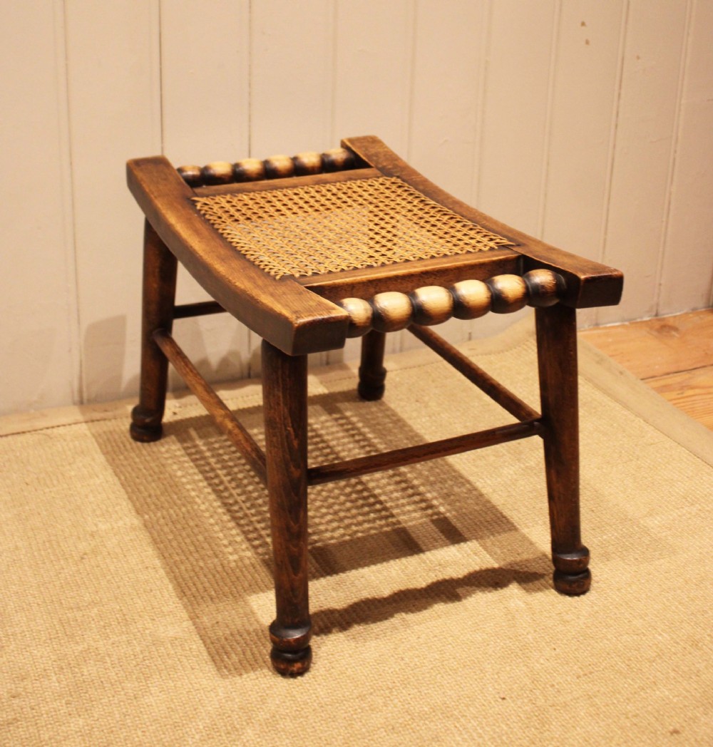An antique warm brown beechwood stool featuring a woven cane seat, bobbin-turned side handles, and splayed turned legs. View VI