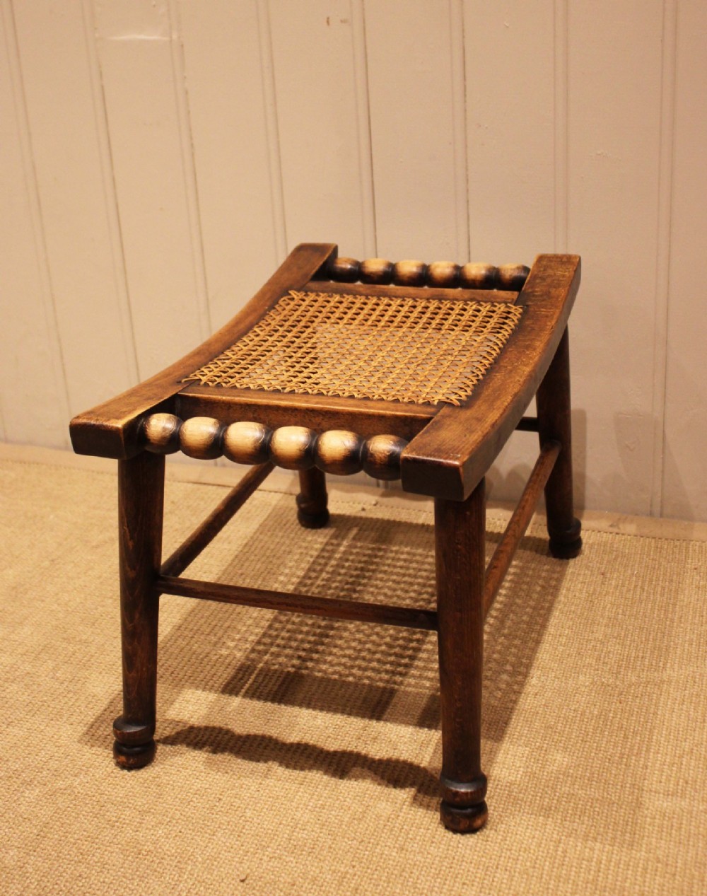 An antique warm brown beechwood stool featuring a woven cane seat, bobbin-turned side handles, and splayed turned legs. View VII