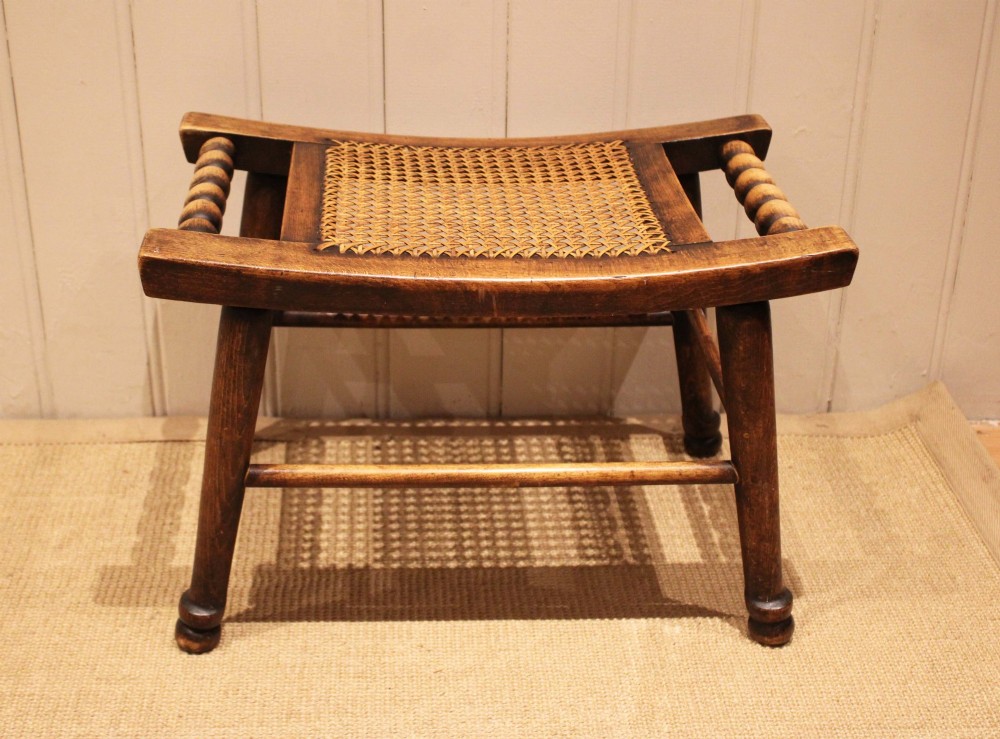 An antique warm brown beechwood stool featuring a woven cane seat, bobbin-turned side handles, and splayed turned legs. View IX