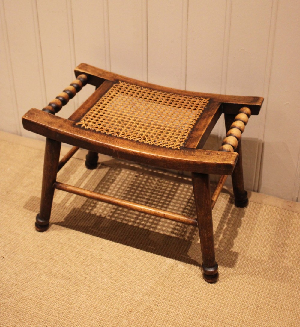 An antique warm brown beechwood stool featuring a woven cane seat, bobbin-turned side handles, and splayed turned legs. View X