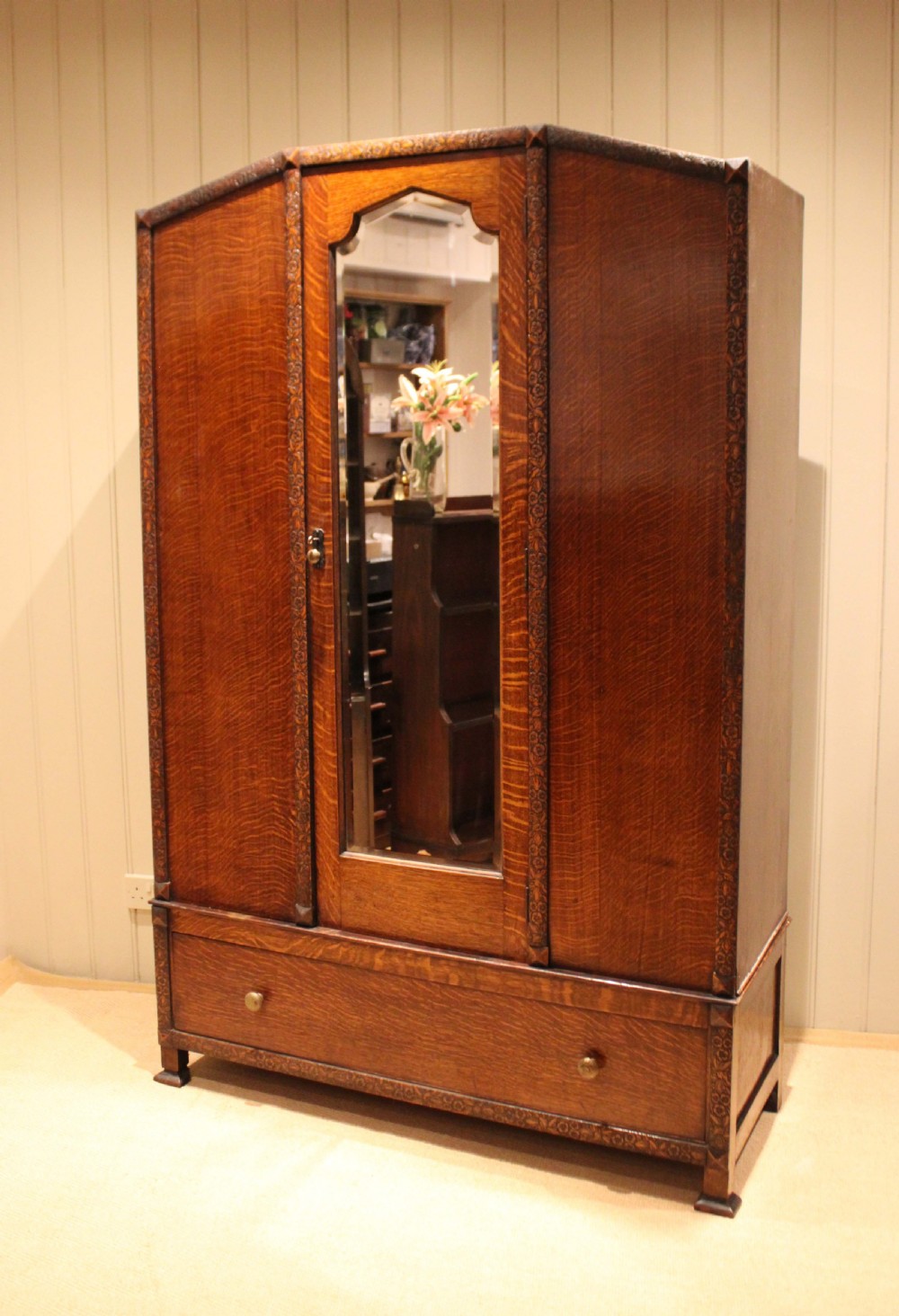 An early twentieth-century warm brown oak wardrobe with a central arched mirror door, carved foliate border mouldings, and a single lower drawer. View X