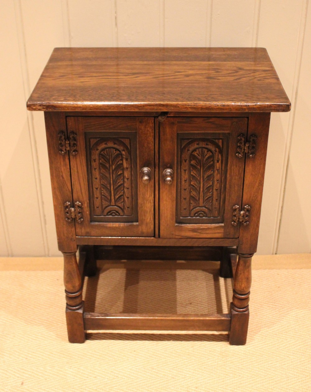 A small dark oak side cabinet featuring double carved panel doors, iron hinges, turned legs, and lower wooden stretchers. View III
