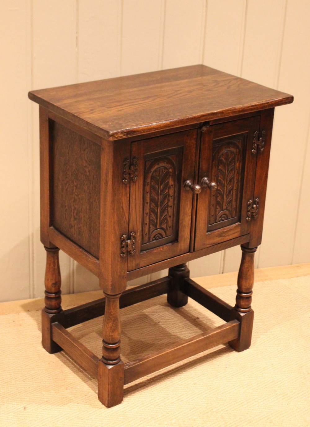 A small dark oak side cabinet featuring double carved panel doors, iron hinges, turned legs, and lower wooden stretchers. View XII