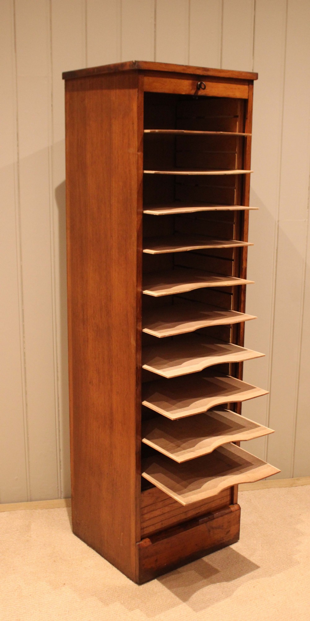 A tall, slender oak filing cabinet featuring a flexible roll-down tambour front, a metal cup handle, a keyhole at the top, and a solid plinth base. View VIII