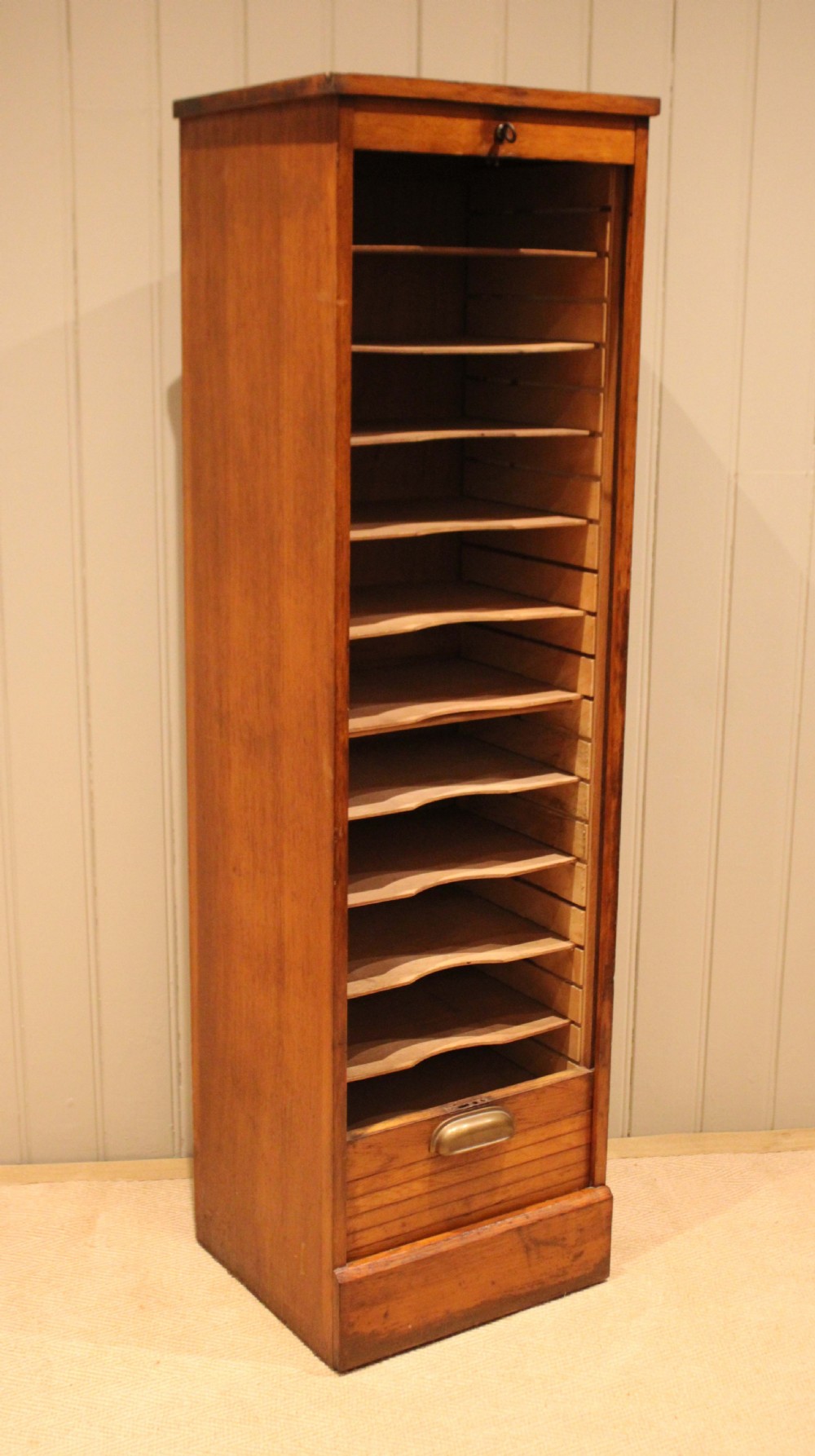 A tall, slender oak filing cabinet featuring a flexible roll-down tambour front, a metal cup handle, a keyhole at the top, and a solid plinth base. View X