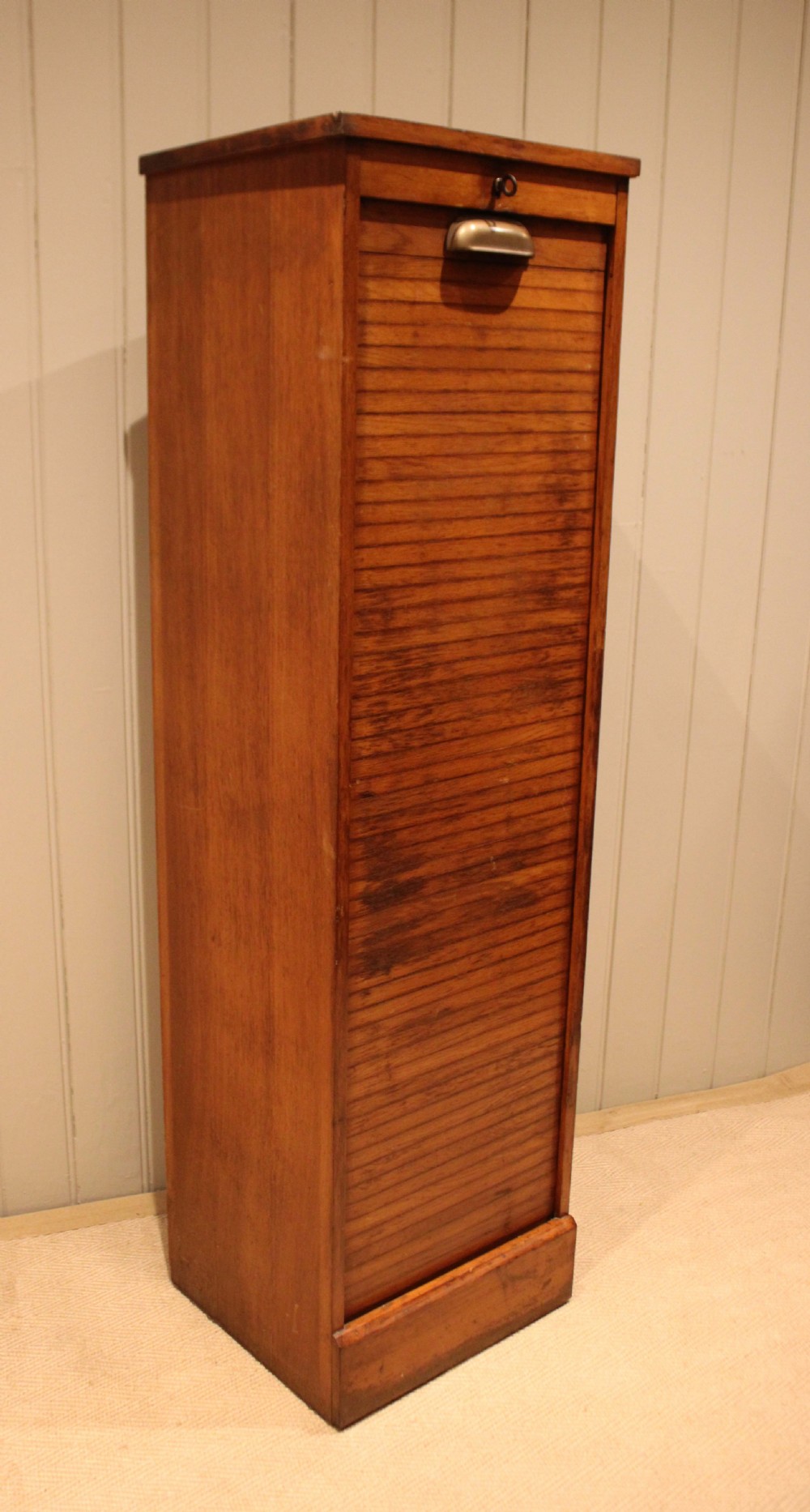 A tall, slender oak filing cabinet featuring a flexible roll-down tambour front, a metal cup handle, a keyhole at the top, and a solid plinth base. View XI
