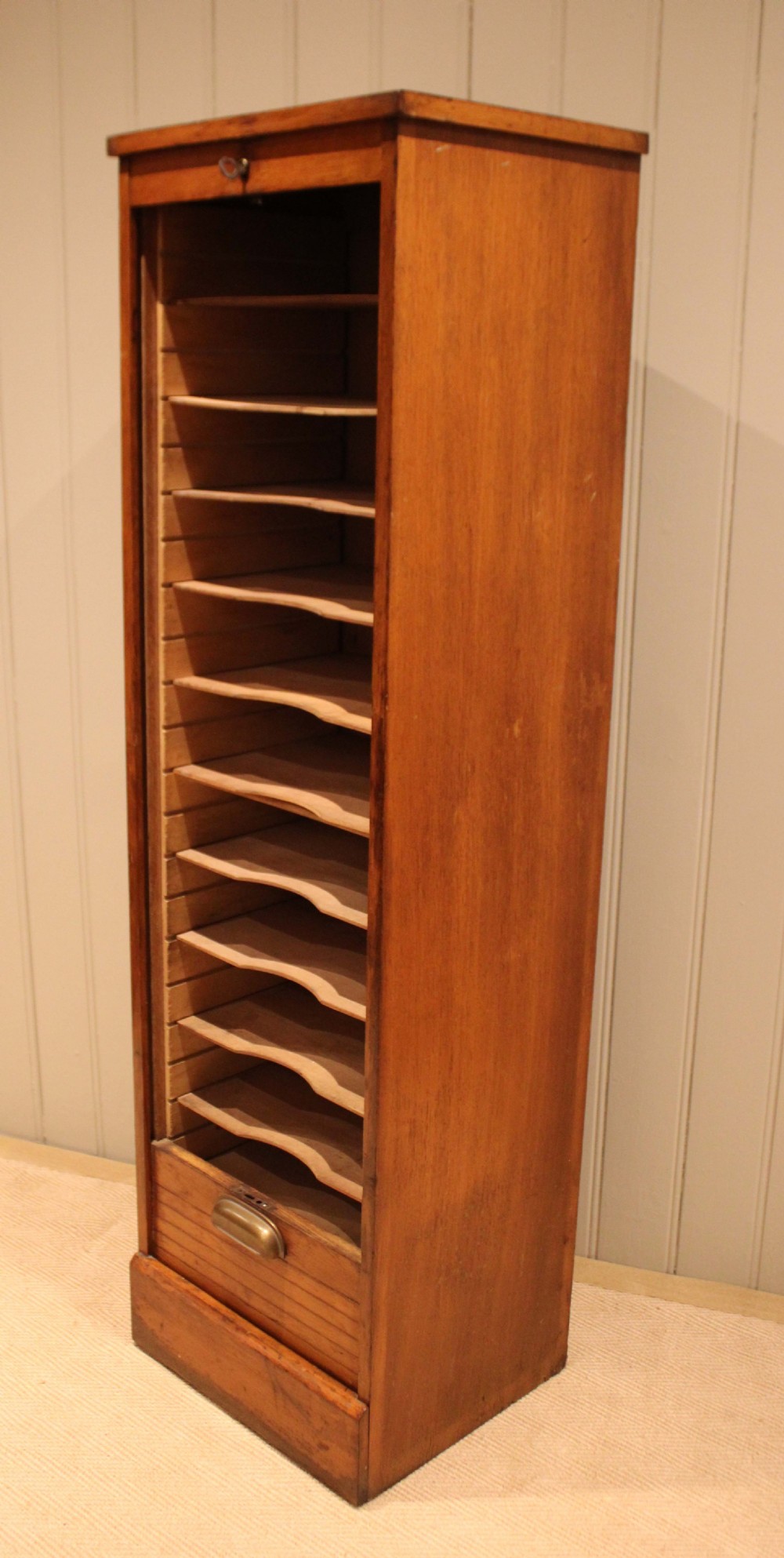 A tall, slender oak filing cabinet featuring a flexible roll-down tambour front, a metal cup handle, a keyhole at the top, and a solid plinth base. View XII