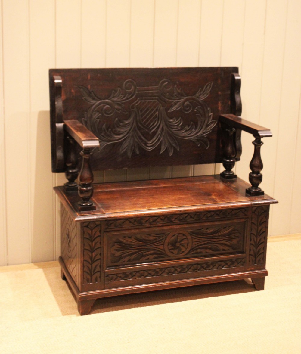 A carved dark oak monks bench featuring a relief-carved backrest, turned arm supports, and a deep box chest storage base.