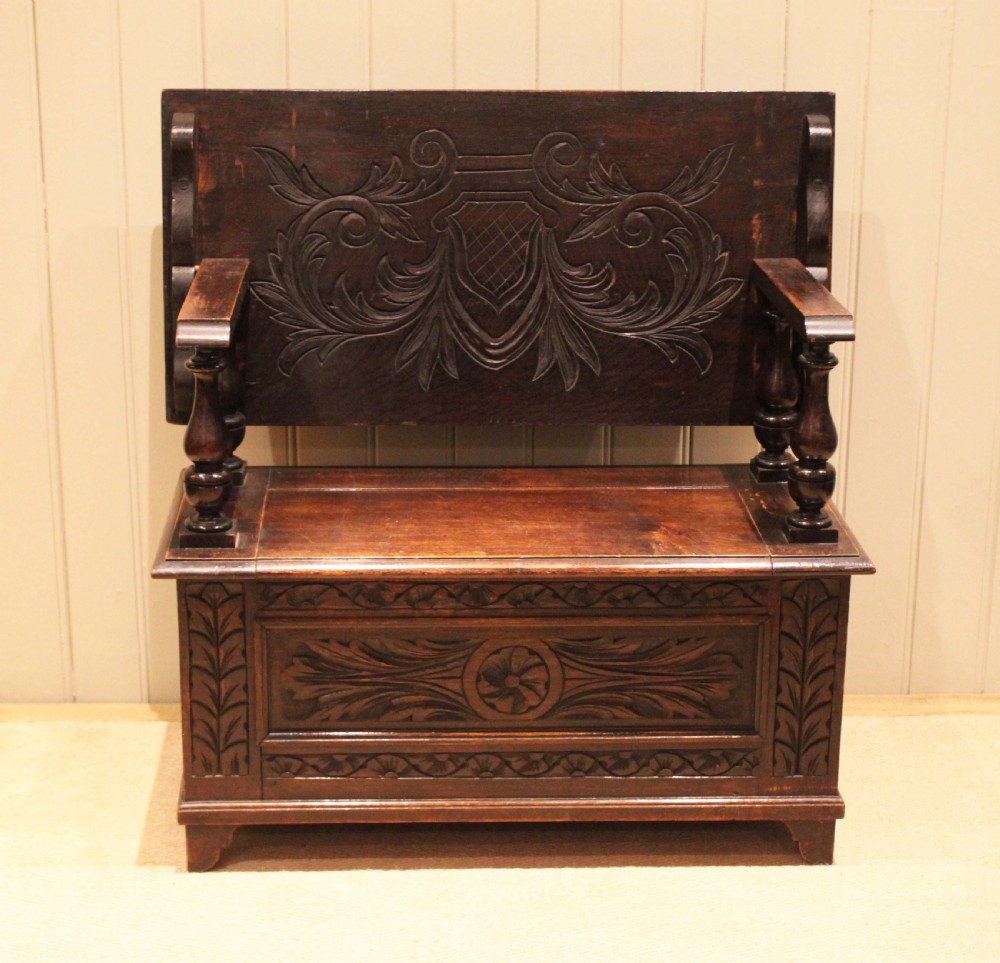 A carved dark oak monks bench featuring a relief-carved backrest, turned arm supports, and a deep box chest storage base. View II