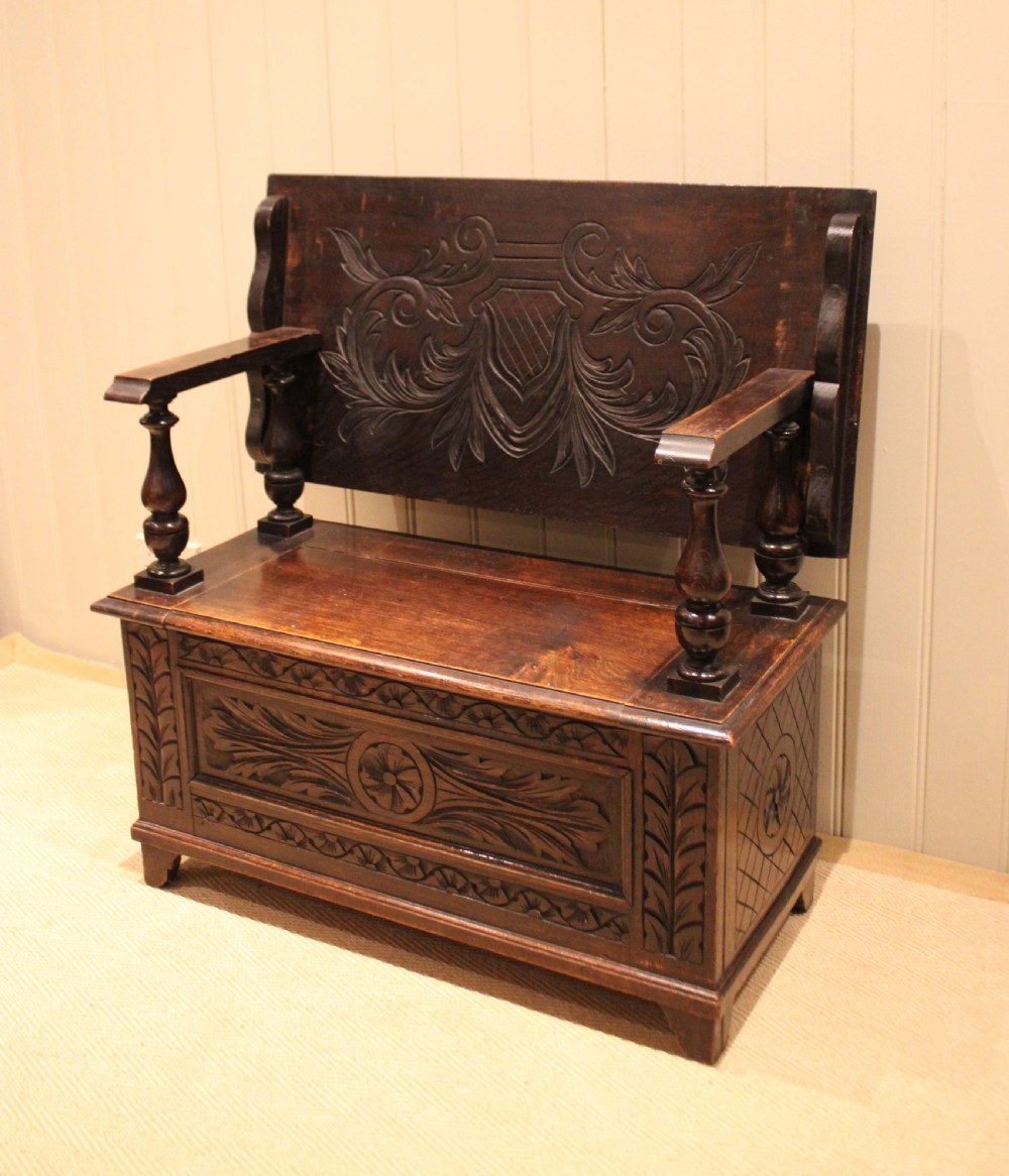 A carved dark oak monks bench featuring a relief-carved backrest, turned arm supports, and a deep box chest storage base. View III