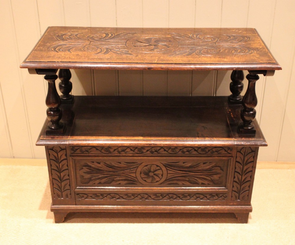 A carved dark oak monks bench featuring a relief-carved backrest, turned arm supports, and a deep box chest storage base. View IV