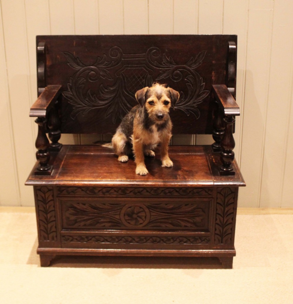 A carved dark oak monks bench featuring a relief-carved backrest, turned arm supports, and a deep box chest storage base. View V