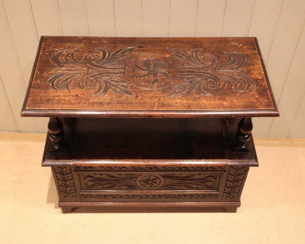 A carved dark oak monks bench featuring a relief-carved backrest, turned arm supports, and a deep box chest storage base. View VI