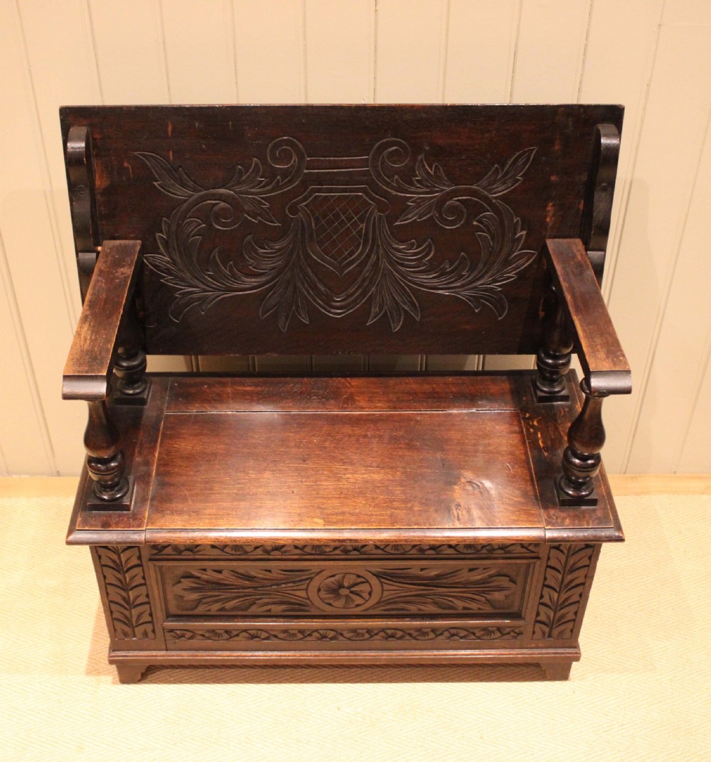 A carved dark oak monks bench featuring a relief-carved backrest, turned arm supports, and a deep box chest storage base. View VII