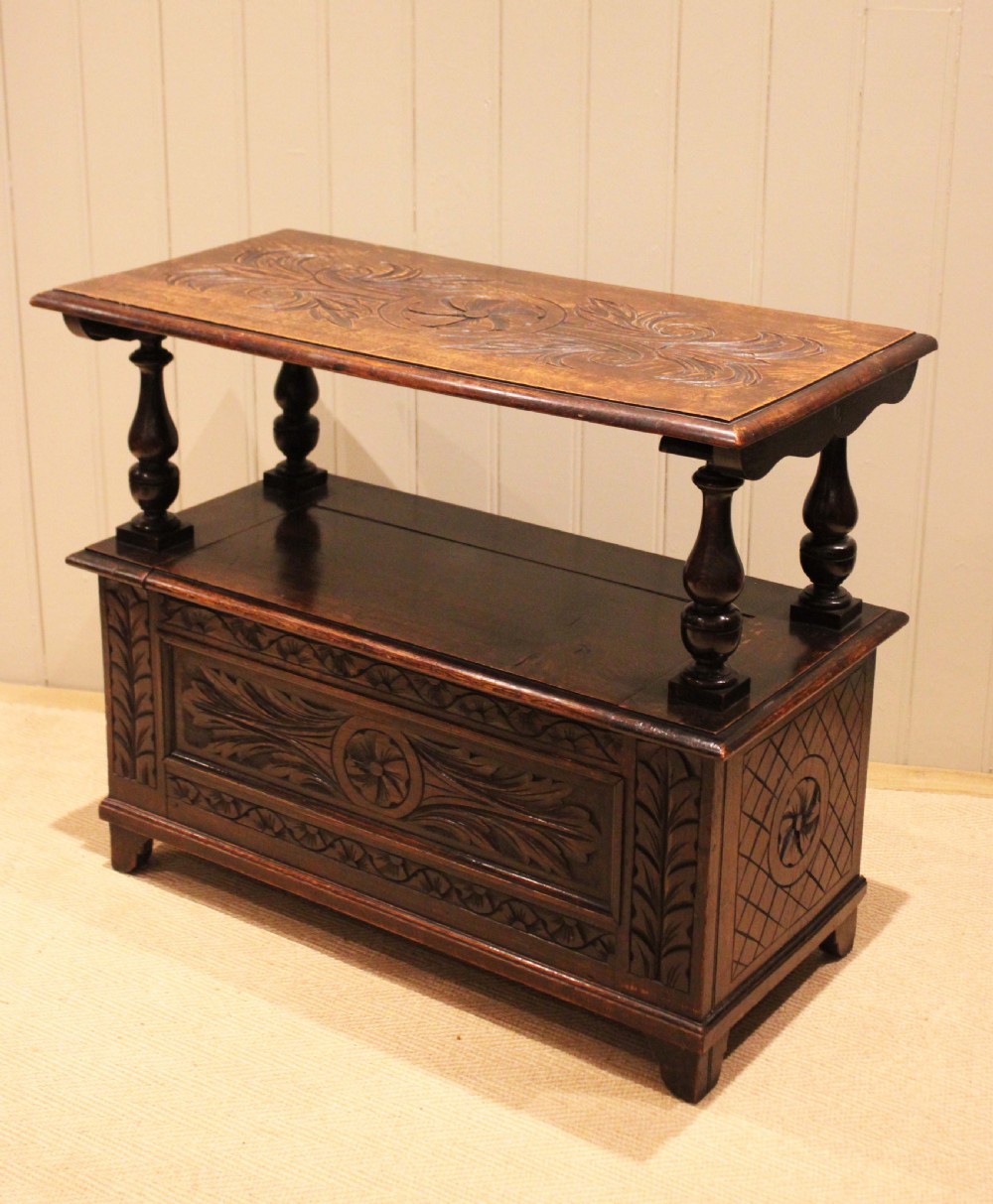 A carved dark oak monks bench featuring a relief-carved backrest, turned arm supports, and a deep box chest storage base. View VIII