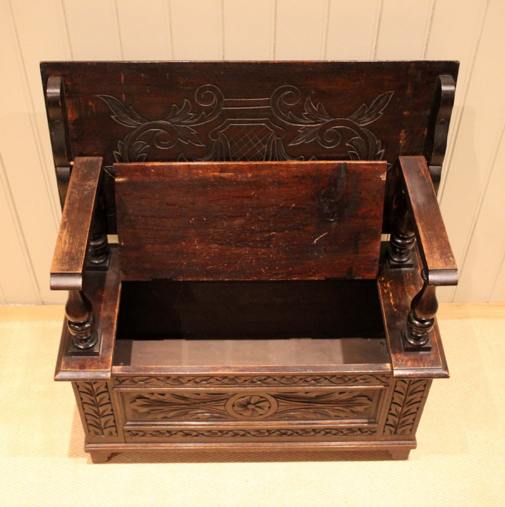 A carved dark oak monks bench featuring a relief-carved backrest, turned arm supports, and a deep box chest storage base. View IX