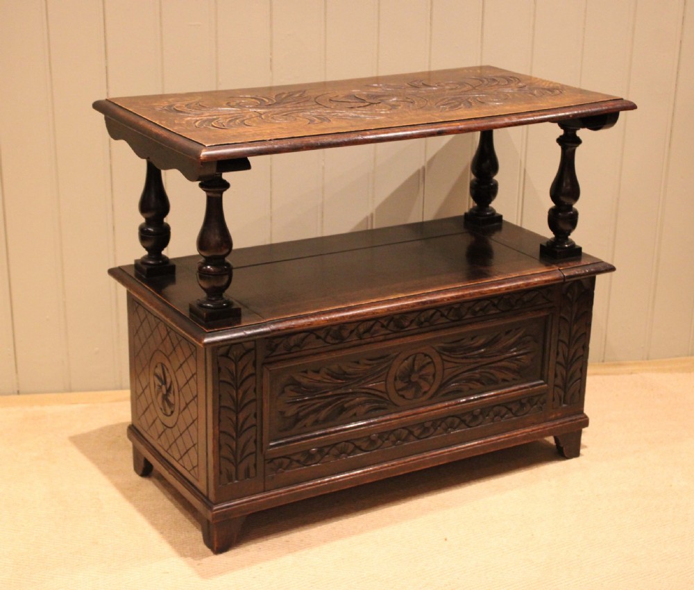 A carved dark oak monks bench featuring a relief-carved backrest, turned arm supports, and a deep box chest storage base. View X