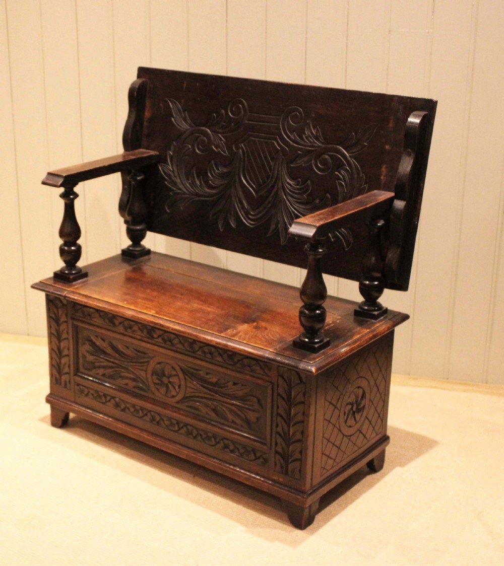 A carved dark oak monks bench featuring a relief-carved backrest, turned arm supports, and a deep box chest storage base. View XI