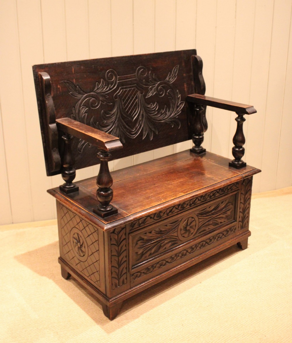 A carved dark oak monks bench featuring a relief-carved backrest, turned arm supports, and a deep box chest storage base. View XII